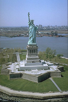 Luftaufnahme der Freiheitsstatue, die auf einer Insel steht, umgeben von Wasser, bei klarem Himmel. Die Statue h&auml;lt eine Fackel in ihrer erhobenen rechten Hand und ein Buch in der linken Hand.