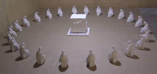 Installation eines k&uuml;nstlerischen Arrangements mit zweiundzwanzig gleichartig gekleideten Figuren, die symmetrisch um einen zentralen, leuchtenden Kubus positioniert sind, in einem Raum mit sandfarbenem Boden und W&auml;nden.