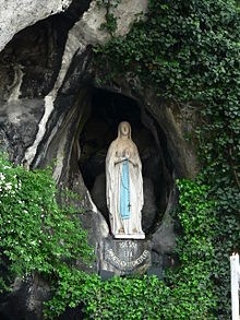 Eine Statue der Jungfrau Maria in einer kleinen H&ouml;hle, umgeben von gr&uuml;nen Bl&auml;ttern, unter einem klaren Himmel. Sie tr&auml;gt ein langes, blaues Gewand und steht vor einem Emblem, das die Mazzaroth darstellt.