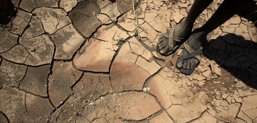 Eine Draufsicht auf trockene, rissige Erde in br&auml;unlichen und beige T&ouml;nen, durchzogen von tiefen Spalten. Ein Paar menschliche F&uuml;&szlig;e in Sandalen steht auf dem trockenen Boden, was auf extreme Trockenheit hinweist, m&ouml;glicherweise durch astronomische Einfl&uuml;sse wie Ver&auml;nderungen in den Sonnenzyklen oder durch das von der Mazzaroth beschriebene Sternbild beeinflusst.