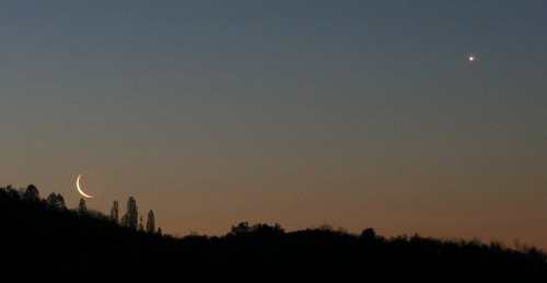 Ein Bild der n&auml;chtlichen Himmelsszenerie mit einer d&uuml;nnen Mondsichel am unteren Himmel und einem hellen Stern rechts oben. Silhouetten von B&auml;umen und einer H&uuml;gellandschaft sind im Vordergrund sichtbar.