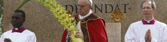 Drei christliche Geistliche w&auml;hrend einer Zeremonie im Freien; der mittlere tr&auml;gt rote Gew&auml;nder und h&auml;lt gro&szlig;e Palmzweige, flankiert von zwei weiteren in wei&szlig;en und purpurnen Gew&auml;ndern. Ein goldener Stuhl und eine Inschrift im Hintergrund sind teilweise sichtbar. Interner Link zu einem Artikel auf dieser Website.