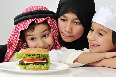 Drei Kinder unterschiedlichen Alters, gekleidet in traditionelle arabische Kopfbedeckungen, betrachten l&auml;chelnd einen Hamburger auf einem wei&szlig;en Teller vor ihnen. Der j&uuml;ngste Junge mit einem rot-wei&szlig;en Kopftuch neigt seinen Kopf zum Burger, w&auml;hrend ein M&auml;dchen mit einem schwarzen Hijab und ein weiterer Junge in einem wei&szlig;en Kopftuch seitlich zusehen.