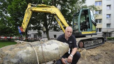 Ein Mann sitzt neben einem gro&szlig;en, ausgegrabenen Objekt, m&ouml;glicherweise eine alte Bombe, auf einer Baustelle. Im Hintergrund ist ein gelber Bagger zu sehen.