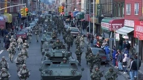 Milit&auml;rfahrzeuge und Soldaten marschieren in einer st&auml;dtischen Stra&szlig;e, umgeben von Gesch&auml;ften und Zuschauern am Stra&szlig;enrand.