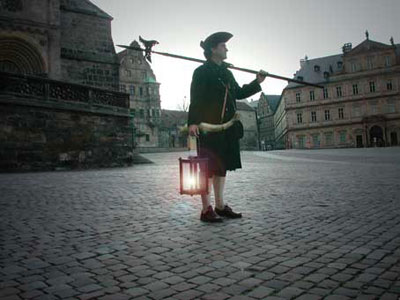 Ein Nachtw&auml;chter in historischer Kleidung steht auf einem gepflasterten Platz und h&auml;lt eine Laterne sowie eine lange Stange. Er ist von alten europ&auml;ischen Geb&auml;uden umgeben, es d&auml;mmert.