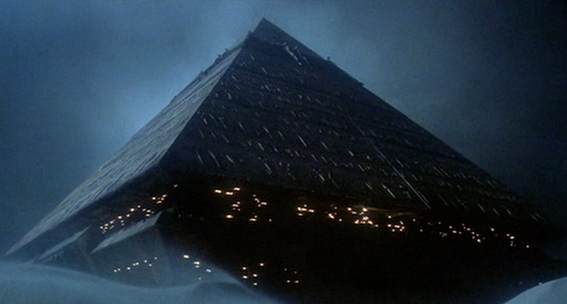 Eine Ansicht einer gro&szlig;en, pyramidalf&ouml;rmigen Struktur in einer n&auml;chtlichen Meereslandschaft, beleuchtet durch zahlreiche Lichter, die durch die Wolken und Dunst schemenhaft sichtbar sind.