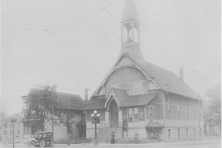 Historisches Foto einer Kirche mit spitzem Turm und angeschlossenem Nebengeb&auml;ude, im Vordergrund ein geparktes altes Auto und zwei Personen, die vor der Kirche stehen.
