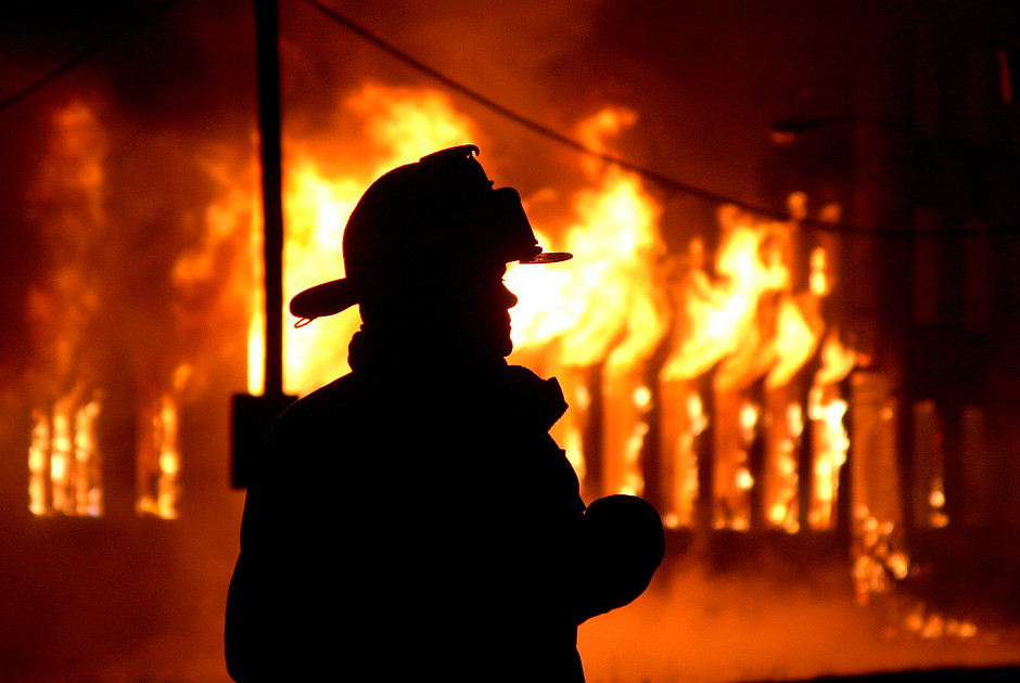 Silhouette eines Feuerwehrmanns vor einem gro&szlig;en Feuer bei Nacht. Deutliche Flammen erhellen den Hintergrund, w&auml;hrend der Feuerwehrmann nachdenklich zur Seite blickt.
