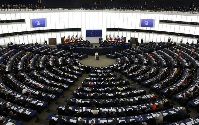 Innenansicht des Europ&auml;ischen Parlaments w&auml;hrend einer Sitzung. Die Abgeordneten sitzen in einem halbkreisf&ouml;rmigen Saal mit mehreren Reihen und h&ouml;ren einem Redner zu. &Uuml;berall sind Flaggen der Mitgliedsstaaten sowie das Flagge der Europ&auml;ischen Union zu sehen.