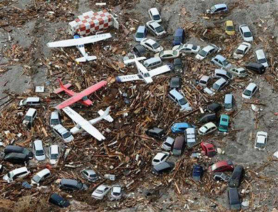 Verschiedene Modellautos und -flugzeuge sind auf einem Boden mit zahlreichen &Auml;sten und Schmutz verstreut. Eine Tischdecke mit Karomuster liegt teilweise sichtbar am Rand.