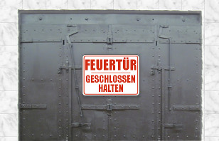 Ein Bild, das eine gro&szlig;e, metallische Sicherheitst&uuml;r zeigt, mit einem roten Schild in der Mitte, auf dem &bdquo;Feuert&uuml;r geschlossen halten&ldquo; steht. Die T&uuml;r weist zahlreiche Schrauben und Verst&auml;rkungen auf und ist gegen eine gemauerte Wand montiert.