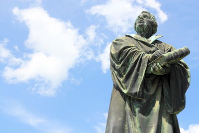 Bronzestatue eines stehenden Mannes vor einem klaren, blauen Himmel, der gewandet ist und seine H&auml;nde vor sich verschr&auml;nkt h&auml;lt.