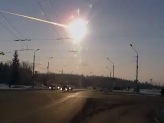 Ein Bild zeigt eine st&auml;dtische Stra&szlig;enszene bei Sonnenuntergang mit einem hellen Meteor, der am klaren Himmel sichtbar ist. Autos beleuchten die Stra&szlig;e und Laternen s&auml;umen die Seiten.