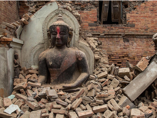 Eine Buddhastatue, eingebettet in Ziegeltr&uuml;mmer, vor dem Hintergrund einer teils zerst&ouml;rten Backsteinmauer und einem verfallenen Geb&auml;ude. Die Statue vermittelt Ruhe trotz der umgebenden Zerst&ouml;rung.