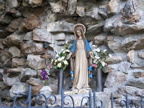 Statue einer religi&ouml;sen Figur, vermutlich der Jungfrau Maria, umgeben von farbigen Blumen und einem kleinen Kreuz, eingebettet in eine rustikale Steinwand.