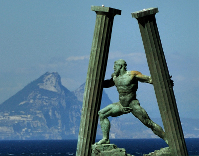 Eine Bronzestatue eines muskul&ouml;sen Mannes, der sich kraftvoll zwischen zwei hohen Steins&auml;ulen durchbewegt, im Hintergrund der Blick auf einen entfernten Berg &uuml;ber dem Meer.
