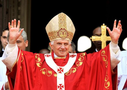 Ein &auml;lterer Mann in religi&ouml;ser Kleidung mit einer Mitra und einem roten Gewand hebt die H&auml;nde vor einer Menschenmenge.