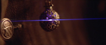 Ein kugelf&ouml;rmiger Anh&auml;nger mit filigranen Metallverzierungen h&auml;ngt von einer Kette, beleuchtet von einem schmalen, horizontalen blauen Lichtstrahl in einer dunklen Umgebung.