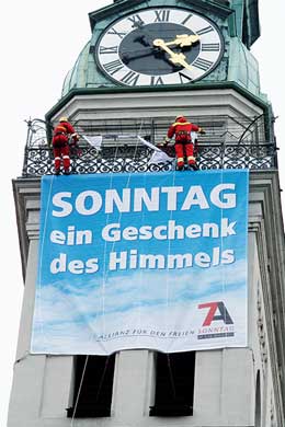 Ein Kirchturm mit einer gro&szlig;en Uhr zeigt die Zeit an, w&auml;hrend oben zwei Figuren, die wie Weihnachtsm&auml;nner aussehen, t&auml;tig sind. Darunter h&auml;ngt ein gro&szlig;es blaues Banner mit der Aufschrift &bdquo;SONNTAG ein Geschenk des Himmels&ldquo;.