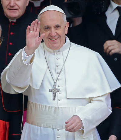 Ein Mann in religi&ouml;ser wei&szlig;en Kleidung und einer gro&szlig;en Kruzifix-Kette l&auml;chelt freundlich und winkt mit seiner Hand. Er steht vor einem Hintergrund von Personen in dunklen Gew&auml;ndern.