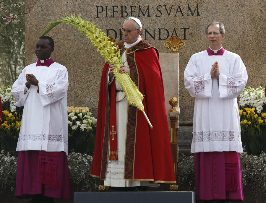 Ein religi&ouml;ses Ritual w&auml;hrend einer christlichen Zeremonie mit drei Geistlichen in liturgischen Gew&auml;ndern. Ein Geistlicher in der Mitte h&auml;lt eine gro&szlig;e, geschwungene Palme. Im Hintergrund sind bl&uuml;hende Blumen und eine steinerne Inschrift zu sehen.