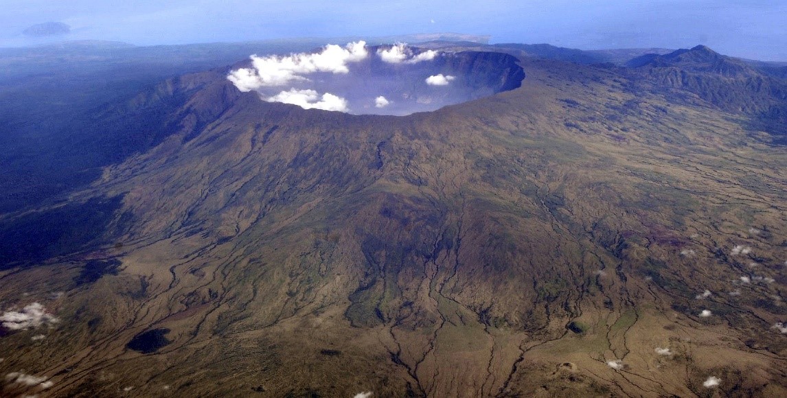 Luftaufnahme eines gro&szlig;en Bergplateaus mit einem prominenten Krater, umgeben von rauen, gr&uuml;nen und braunen Gel&auml;ndestrukturen, unter einem klaren Blauen Himmel mit wei&szlig;en Wolken.