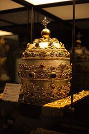 Ein kunstvoll verziertes goldenes Objekt, m&ouml;glicherweise eine Krone oder ein Zeremonialgegenstand, beleuchtet in einer Vitrine. Die Verzierungen umfassen feine Gravuren und eventuell Darstellungen aus der Mazzaroth.