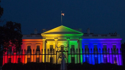Bild zeigt das Wei&szlig;e Haus in der Nacht, beleuchtet mit einem Spektrum von Farben, die sich &uuml;ber den Regenbogen erstrecken, vor einem dunklen Himmel.