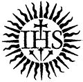 Ein grafisches Symbol mit einem zentralen Monogramm "IHS" umgeben von Strahlen, die einem klassischen Gestirn &auml;hneln. Das Monogramm ist durch einen Kreuzanker gekr&ouml;nt.