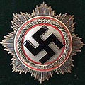 Abzeichen mit einem schwarzen Hakenkreuz in der Mitte, umgeben von einem wei&szlig;en Kreis und einem roten Ring, eingerahmt von einem silbernen Sternenkranz auf dunklem Hintergrund.